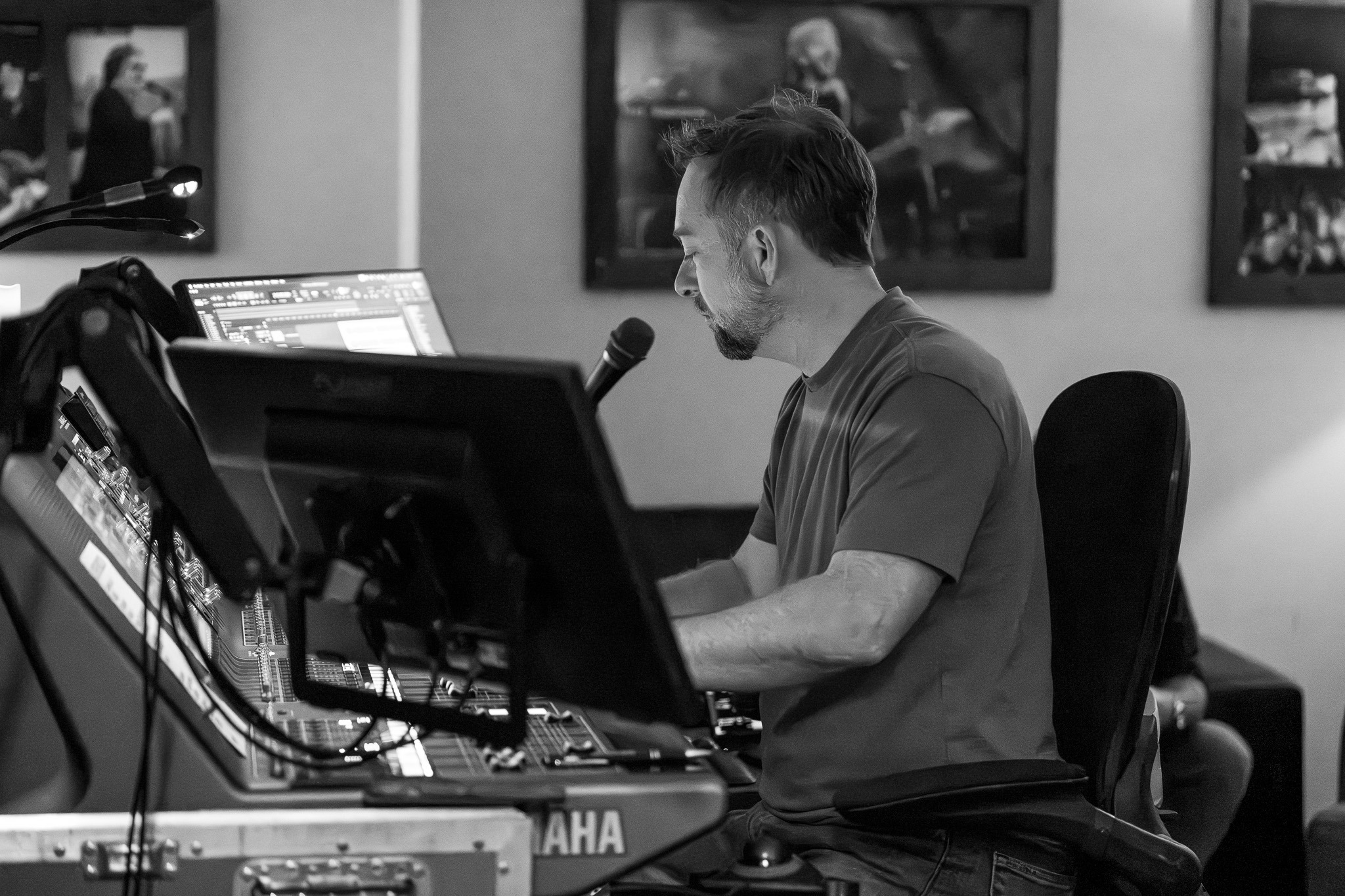 Black and white photo of an audio engineer mixing at a Yamaha digital console, with framed photographs on the wall behind him