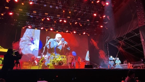 Band on an outdoor festival stage with a large video screen and lighting trusses