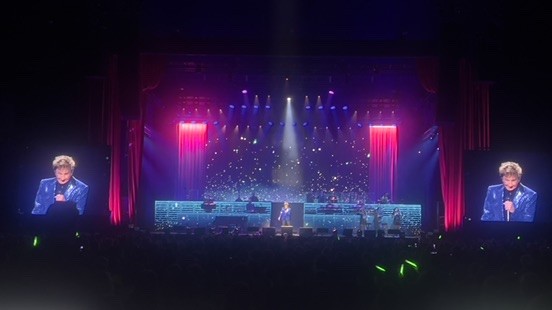 Singer in a blue jacket on a theater stage with red curtains and a video screen on each side