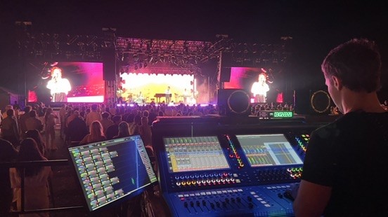 Engineer at a digital console facing an outdoor stage with video screens and a crowd