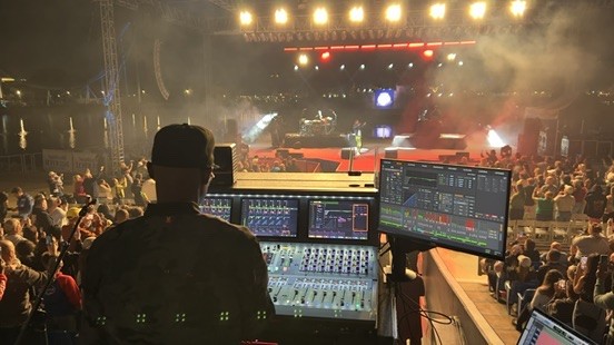 Engineer in a cap at the console with an analyzer screen, facing a waterfront stage and crowd