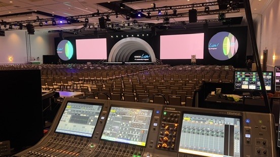 Digital console in the foreground facing a ballroom with a stage and video screens