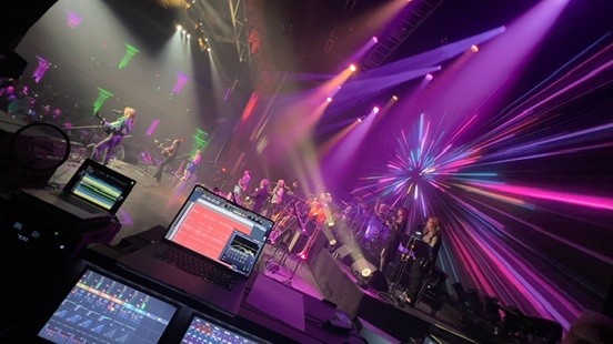 Side-stage view of a band under pink and purple lights with a recording laptop