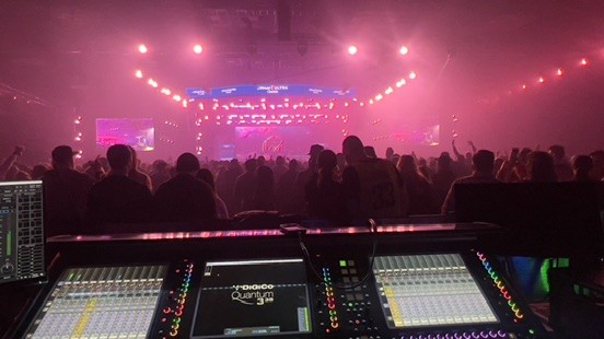 Console and monitor screens at the mix position facing a crowd in an arena washed in pink light