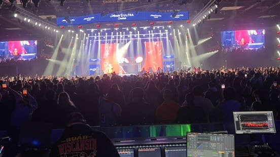 Arena crowd facing a lit stage with video screens, with an analyzer and recording screens at the mix position