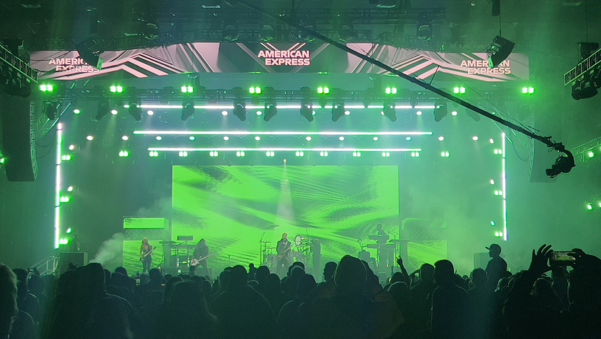 Band on a stage with green lighting, a video wall and a lit truss, seen over the crowd