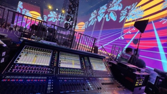 Mixing console on a riser with a video wall showing neon palm trees and a sunset grid