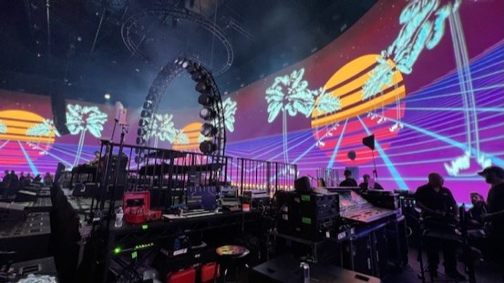 Equipment racks and keyboards at the side of a stage with a curved neon palm-tree video wall